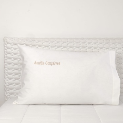 Travesseiro branco com fronhas bordadas com nome, sobre colchão branco com cabeceira acolchoada cinza claro.