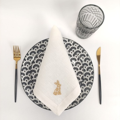 Conjunto de jantar com prato padrão preto e branco, guardanapo branco bordado, talheres dourados e pretos e copo transparente