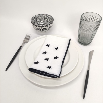 Conjunto de mesa com prato branco, recipiente decorado, copo de vidro e talheres com cabos pretos, guardanapo branco com estrelas pretas