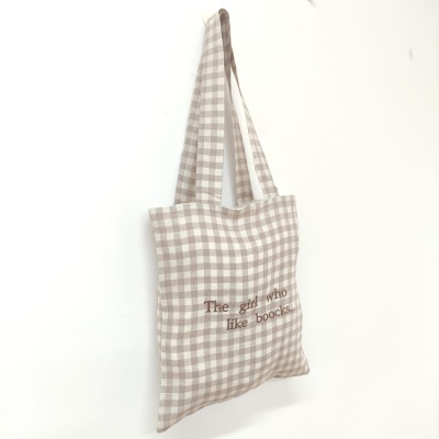 Saco tote bag xadrez bege e branco com frase bordada pendurado numa parede branca