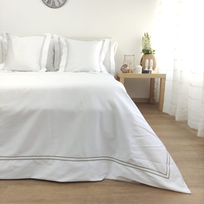 Quarto com cama de roupa de cama branca bordada, cortinas brancas e mesa de madeira com decoração.