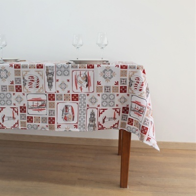 Toalha de mesa retangular estampada com padrões geométricos e figuras sobre mesa de madeira com chão de madeira clara e taças de vidro no fundo