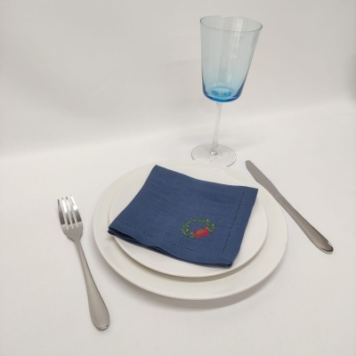 Conjunto de louça branca com guardanapo azul bordado, talheres metálicos e copo azul claro