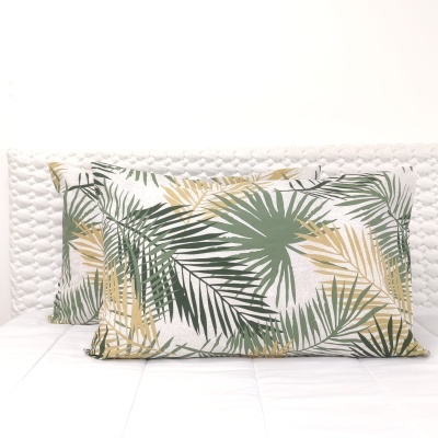 Duas almofadas decorativas com padrão de folhas tropicais verdes e bege sobre fundo branco numa cama com cabeceira branca acolchoada.