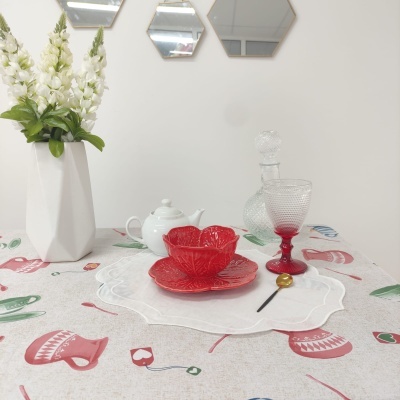 Mesa com toalha estampada, pratos e tigelas vermelhas, jarra de vidro e vaso branco com flores