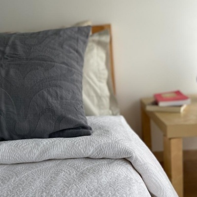 Almofadas cinza e bege sobre cama com colcha branca, mesa de madeira ao fundo com livros