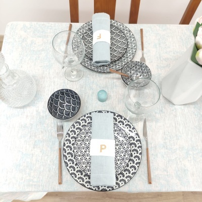 Mesa posta com pratos decorados, guardanapos com letras douradas, e jarra de flores brancas.