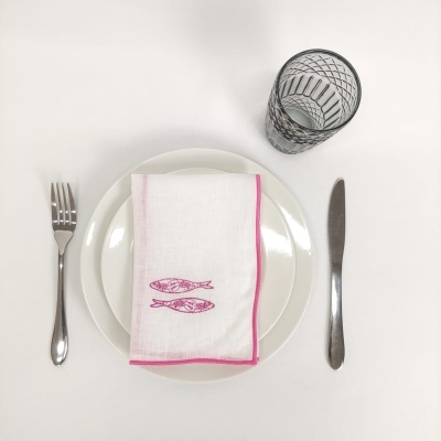 Conjunto de mesa com prato branco, guardanapo rosa e branco com peixes, talheres em inox e copo transparente com padrão