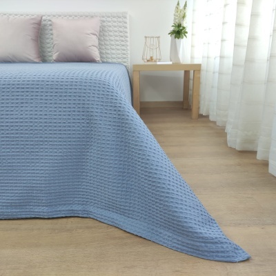 Colcha azul clara com padrão waffle em cama com almofadas cinza claro, mesa de madeira e cortinas brancas