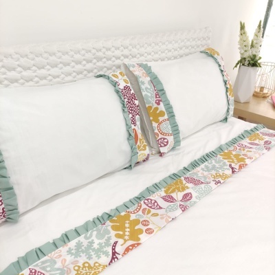 Fronhas brancas com bordas estampadas coloridas e folho verde água num conjunto de cama branco, com mesa de cabeceira com flores ao lado.