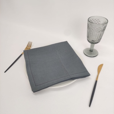 Jogo da mesa com guardanapo cinza, prato branco, talheres dourados e pretos, e copo de vidro cinzento.