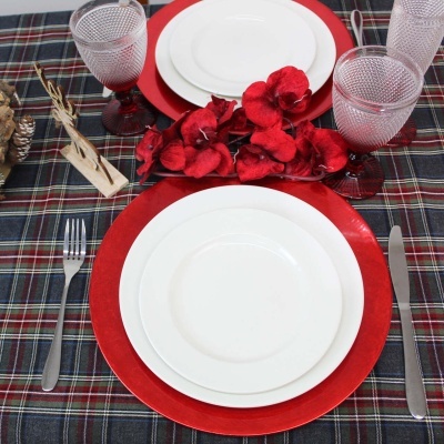 Conjunto de jantar com pratos brancos e bases vermelhas sobre toalha xadrez escura com copos texturizados e flores vermelhas