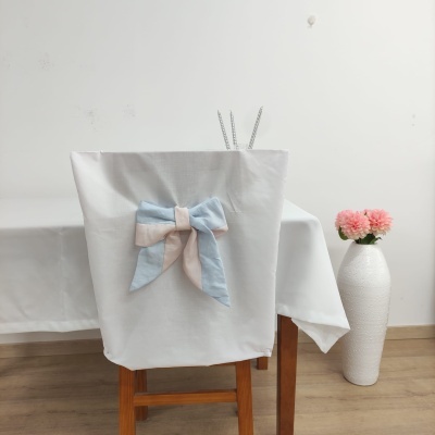 Capa branca para cadeira com laço azul claro e bege numa mesa com toalha branca e vaso com flores cor-de-rosa
