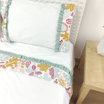 Jogo de cama branco com estampas coloridas e folhos verdes na cama ao lado de mesa de madeira.