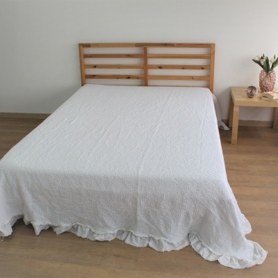 Cama com colcha branca texturizada e cabeceira em madeira clara ao lado de mesa com vaso de flor