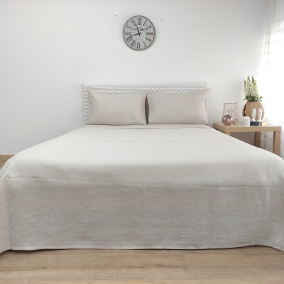 Cama com colcha bege e almofadas, cabeceira branca, mesa de madeira e relógio na parede