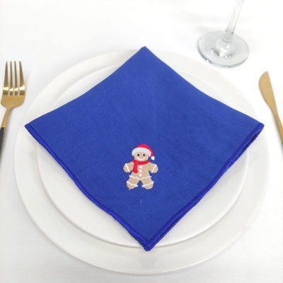 Guardanapo azul com bordado de biscoito de gengibre num prato branco com talheres dourados e copo de vidro.