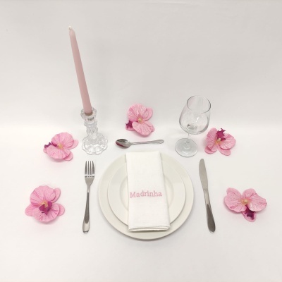 Mesa posta com prato, talheres, guardanapo bordado, copo, vela e flores cor-de-rosa