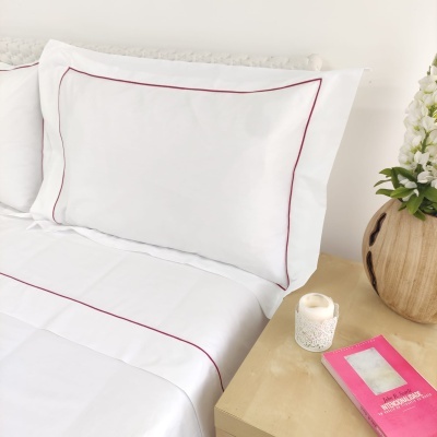 Lençóis brancos com linha vermelha em cama ao lado de mesa com vaso, vela e embalagem rosa