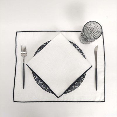 Conjunto de mesa com base branca, prato com padrão escuro, guardanapo branco, talheres pretos e copo transparente