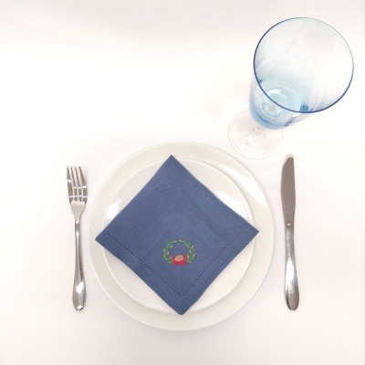 Conjunto de mesa com prato branco, guardanapo azul bordado, talheres metálicos e copo azul