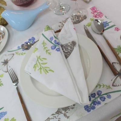 Mesa posta com guardanapo e caminho de mesa florais, prato branco e talheres com cabo de madeira