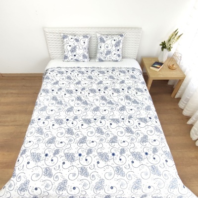 Conjunto de roupa de cama com padrão floral azul e branco numa cama com cabeceira branca e mesa de cabeceira com objetos.