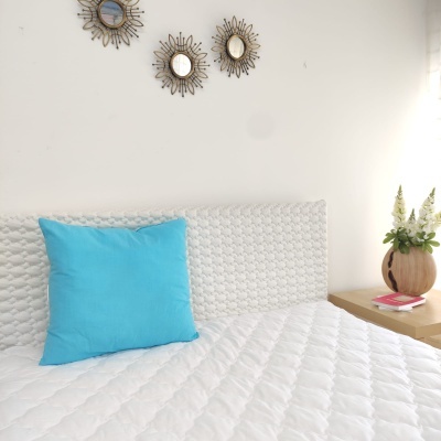 Cama branca com cabeceira estofada, almofada azul, mesa de cabeceira com vaso e livros, espelhos decorativos na parede