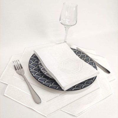 Conjunto de jantar com prato azul decorado, guardanapo branco bordado, talheres metálicos e copo de vidro sobre panos brancos