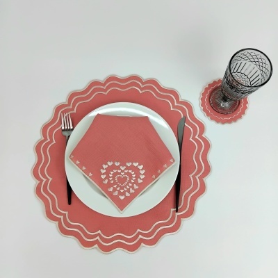 Individual e porta-copo coral e branco com guardanapo rosa decorado, prato branco, talheres pretos e copo transparente com padrão preto