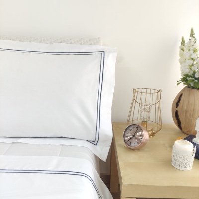 Conjunto de cama branco com risca azul ao lado de mesa de cabeceira com relógio, vela, candeeiro e vaso com flores.