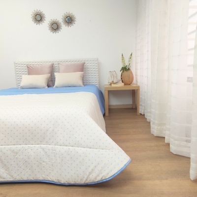 Quarto com cama decorada com edredão branco e azul e almofadas, ao lado mesa de cabeceira e cortinas brancas.
