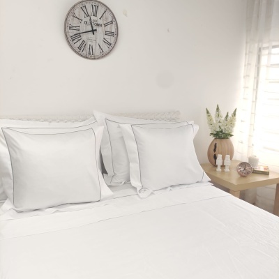 Cama com roupa de cama branca, almofadões, relógio de parede com números romanos e mesa de cabeceira com vaso de flores