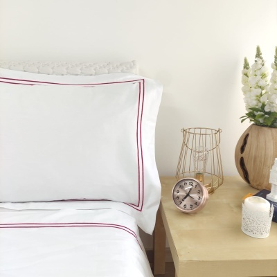 Jogo de cama branco com linhas vermelhas em cama ao lado de mesa amarela com relógio, castiçal, vela e vaso com flores.