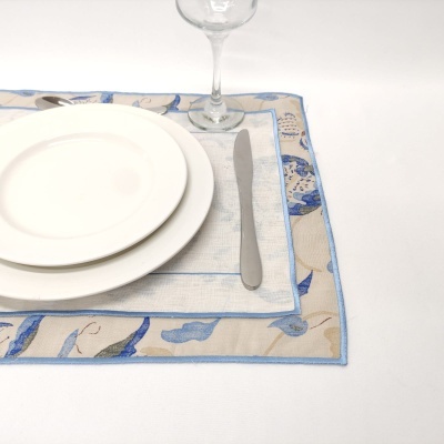 Jogo americano com padrão azul e bege, pratos brancos, talheres e copo de vidro sobre superfície branca.