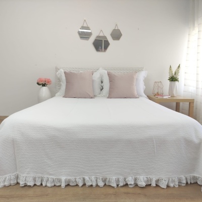 Cama de casal com colcha branca bordada, almofadas castanho claro, cabeceira branca e espelhos geométricos na parede