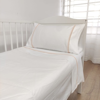 Cama branca com roupa de cama branca e linhas castanho claro, em quarto com cortina branca e chão de madeira clara