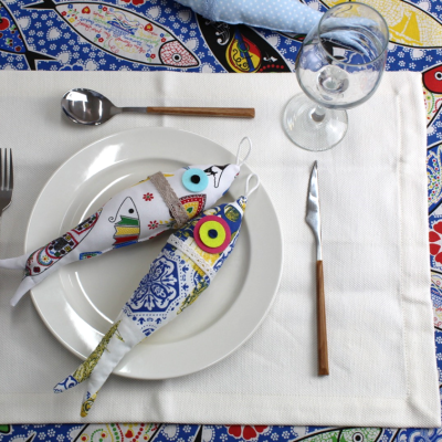 Duas pegas de tecido em forma de peixe com padrões coloridos sobre prato branco, em mesa com toalhas e talheres.