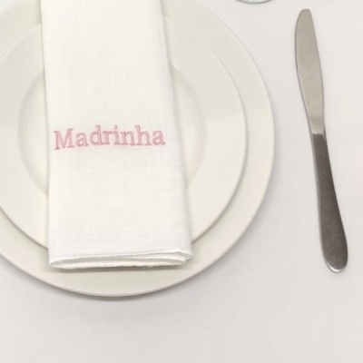 Guardanapo branco com bordado rosa 'Madrinha' sobre pratos brancos e faca ao lado