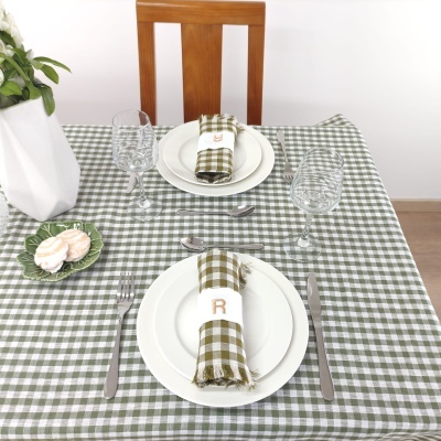 Mesa posta com toalha xadrez verde e branco, pratos brancos, guardanapos com argolas bordadas, talheres metálicos e taças de vidro
