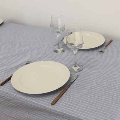 Mesa posta com toalha azul com riscas brancas, pratos brancos, copos de vidro e talheres de cabos castanhos.