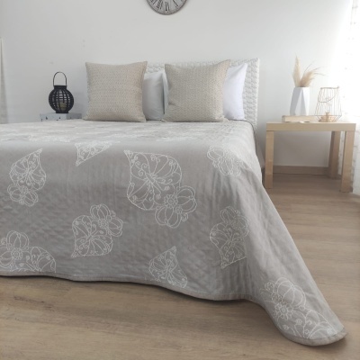 Cama com colcha cinzenta de padrão floral branco, almofadas cinzentas e brancas, relógio de parede e decoração minimalista