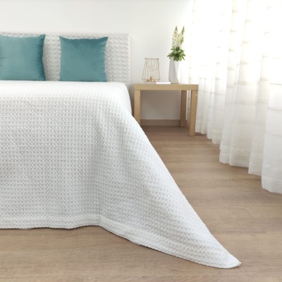 Cama com cobertura branca texturizada, almofadas azuis, mesa de madeira com flores e cortinas brancas translúcidas