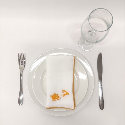 Conjunto de mesa com prato branco, guardanapo branco com bordas amarelas e desenhos de trenó e rena em amarelo, talheres de metal e copo de vidro com água.