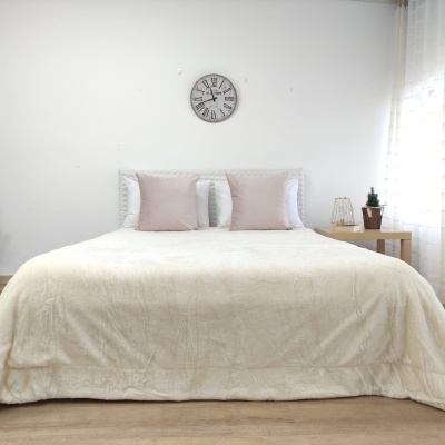 Cama de casal com almofadas e cobertor bege clara num quarto com relógio de parede e mesa de cabeceira