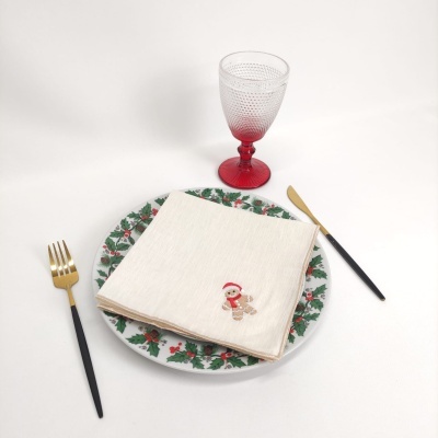 Prato decorado com padrão de azevinho, guardanapo bege com bordado de boneco de gengibre, talheres pretos e dourados e copo com base vermelha.