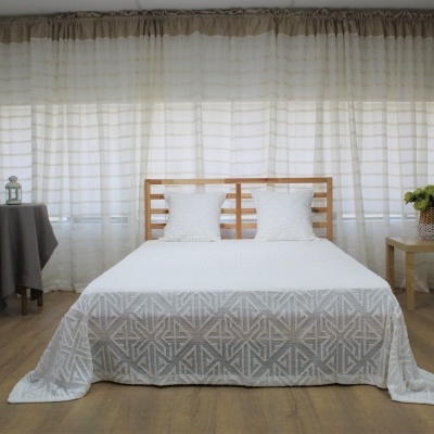 Quarto com cama de madeira, colcha branca com padrões cinza e cortinas brancas