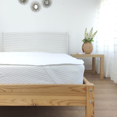 Cama de madeira clara com colchão branco e cabeceira texturada, ao lado mesa com vaso de plantas