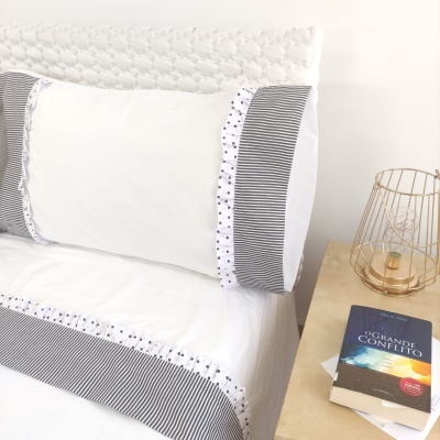 Roupa de cama branca com riscas cinzentas e bolinhas pretas ao lado de mesa com abajur dourado e livro