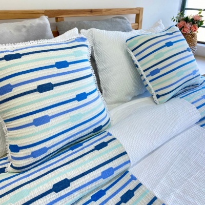 Almofadas e colcha azul, branca e cinza com padrões riscados numa cama com cabeceira de madeira e flores ao fundo.